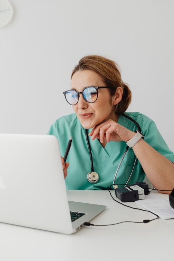 How to Progress from Care Assistant to Senior Carer A healthcare professional wearing scrubs and eyeglasses using a laptop with a stethoscope around the neck.